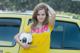 A young woman holding a soccer ball in front of a yellow car.